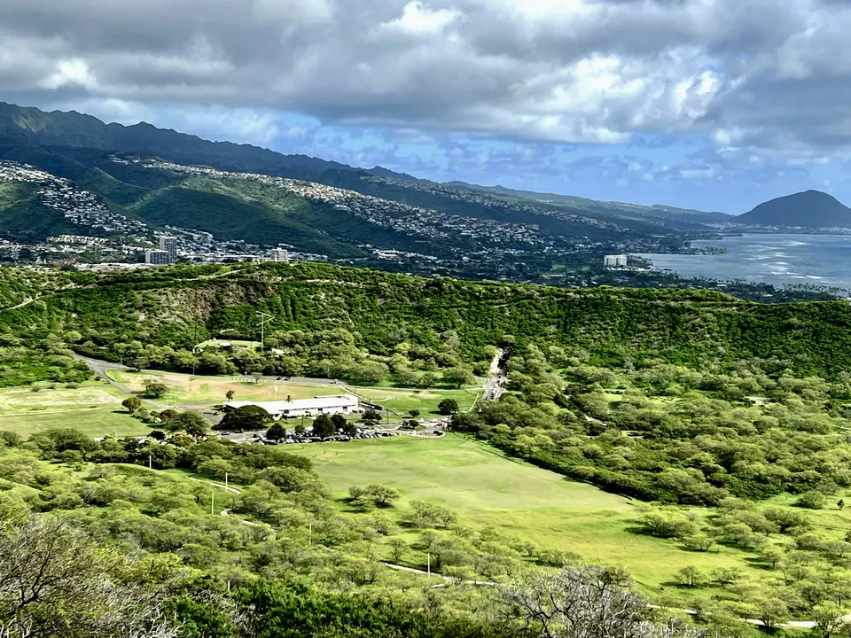 A lush green valley with a golf course overlooks a coastal city, mountains, and the ocean beneath dramatic clouds.