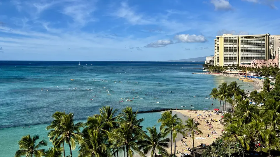 Palm trees frame a crowded sandy beach and turquoise ocean beside high-rise hotels under a blue sky.