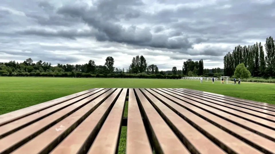 A wooden slatted bench overlooks a green sports field with distant players beneath a cloudy sky.