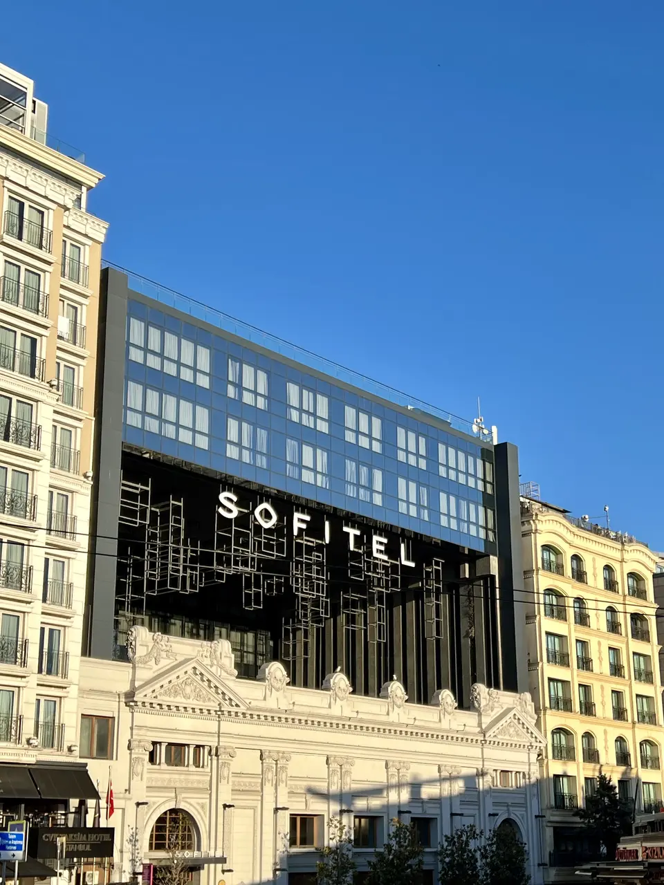 The modern glass-fronted Sofitel building rises behind an ornate white historic facade under a clear blue sky.