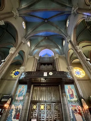 A vaulted church interior features stone columns, colorful stained-glass windows, religious icon panels, and a pipe organ above wooden doors.