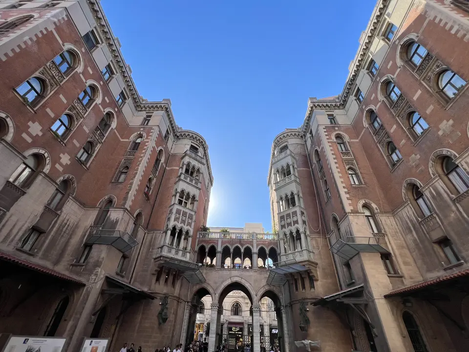 Ornate red brick buildings with arched windows frame a vaulted passageway beneath a clear blue sky.