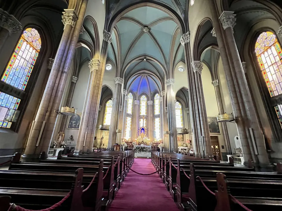 A vaulted church interior with tall stone columns, stained-glass windows, wooden pews, and a red-carpeted central aisle leading to the altar.