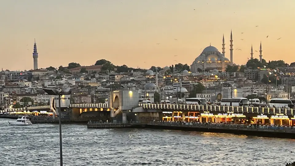 Galata Bridge spans choppy water below a dense hillside of buildings and a domed mosque at sunset.