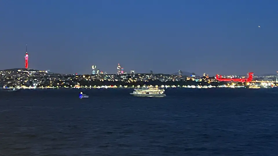 An illuminated suspension bridge and city skyline stretch across dark water at night, with boats in the foreground.