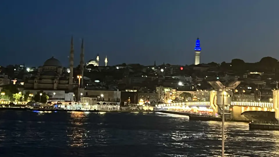 A nighttime waterfront view shows a domed mosque, a blue-lit tower, and illuminated buildings above choppy water.