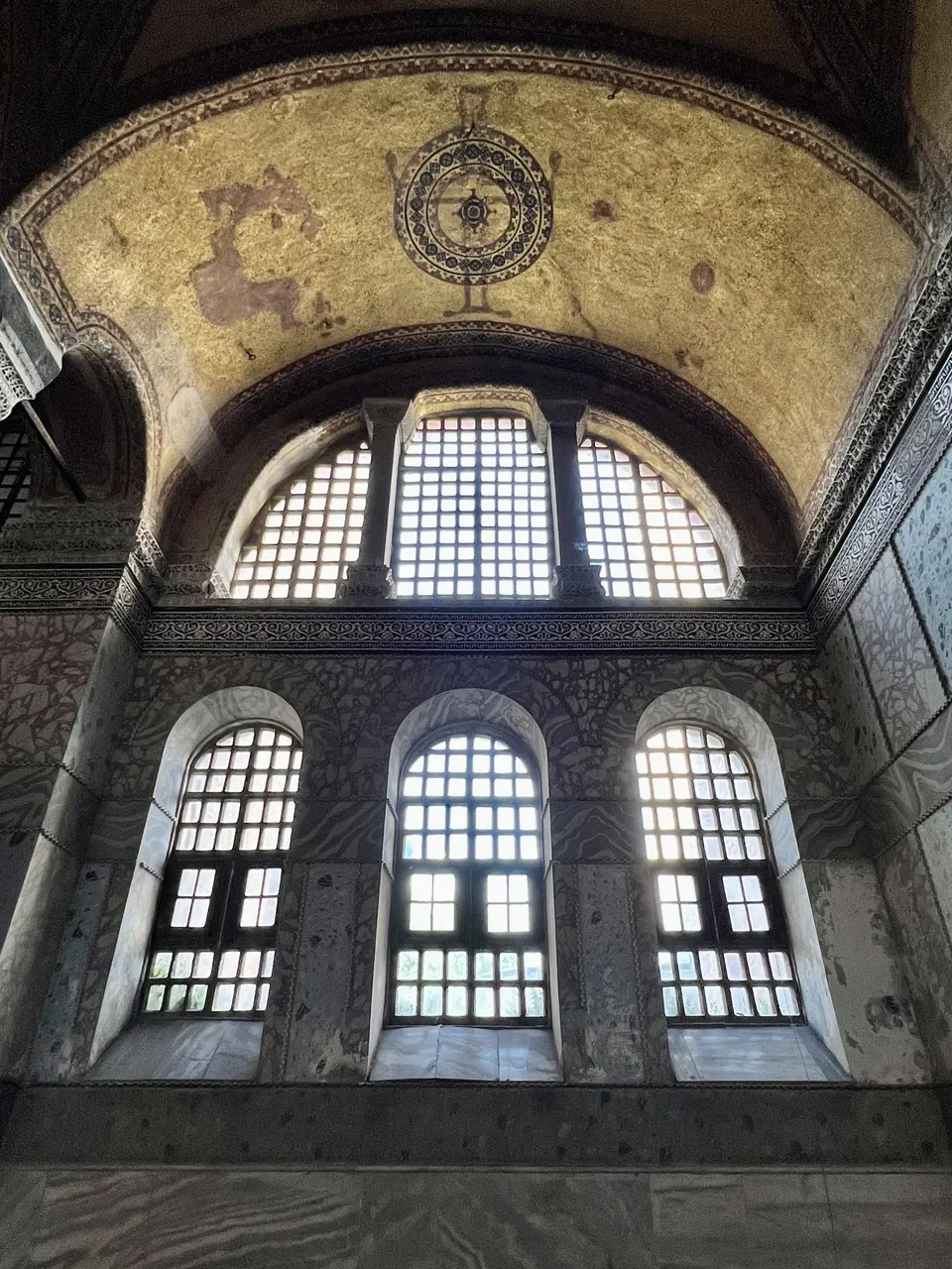 Tall arched windows rise beneath a gilded vaulted ceiling with a circular ornamental mosaic, above veined marble walls.