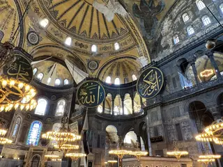 The ornate interior of Hagia Sophia features a painted dome, hanging chandeliers, stained-glass windows, and large Arabic calligraphy medallions.
