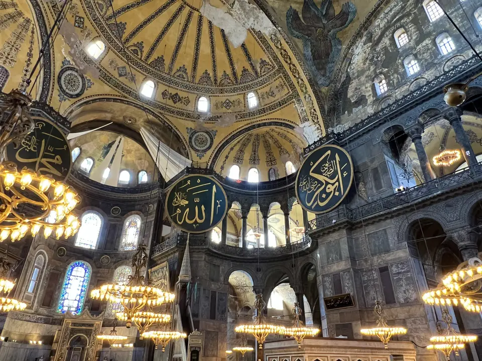 The ornate interior of Hagia Sophia features a painted dome, hanging chandeliers, stained-glass windows, and large Arabic calligraphy medallions.
