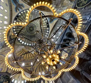 An ornate circular chandelier hangs beneath the richly patterned domes and arches of Hagia Sophia.