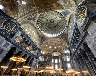 The vast interior of Hagia Sophia rises beneath a decorated central dome, with marble columns, arched galleries, hanging chandeliers, and large Arabic calligraphy medallions.