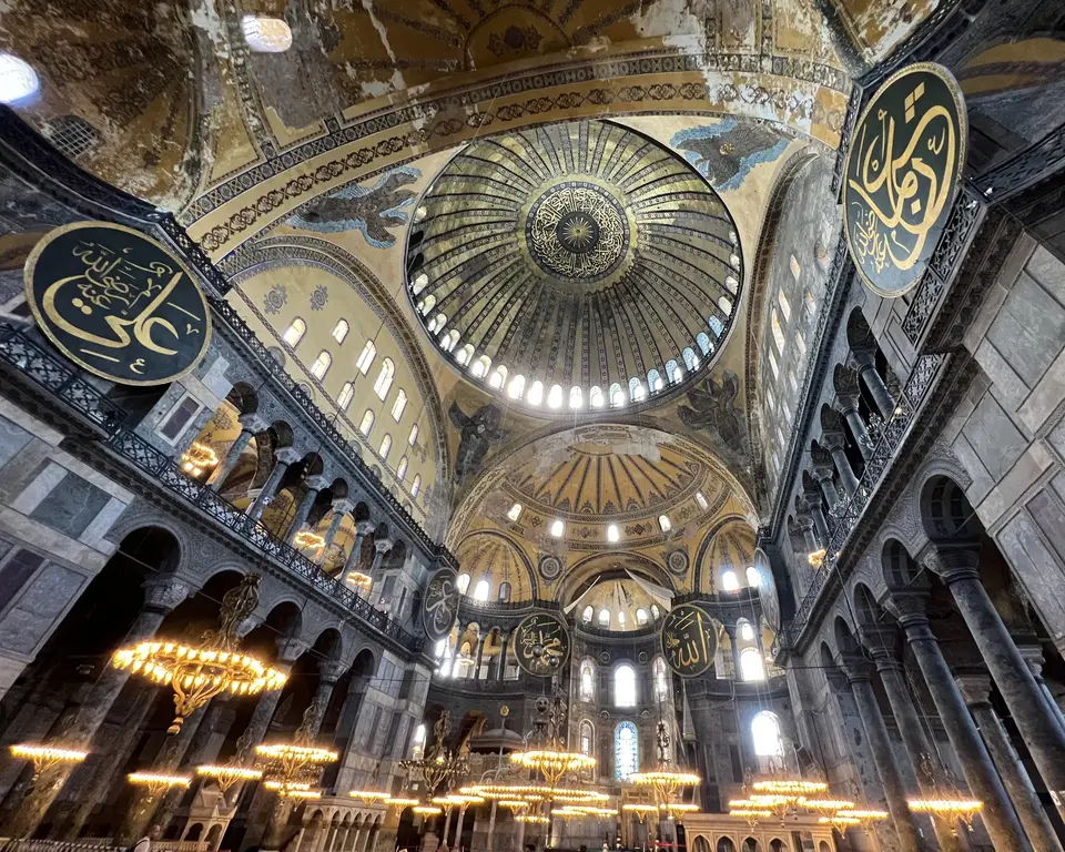 The vast interior of Hagia Sophia rises beneath a decorated central dome, with marble columns, arched galleries, hanging chandeliers, and large Arabic calligraphy medallions.