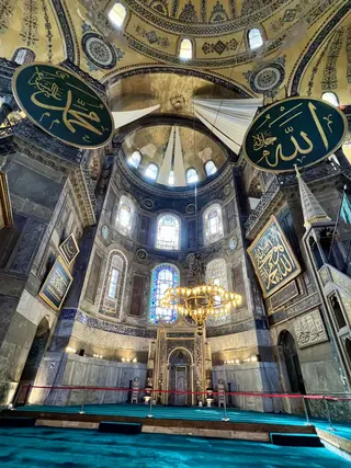 The ornate interior of Hagia Sophia features a painted dome, stained-glass windows, large Arabic calligraphy medallions, and a glowing chandelier.