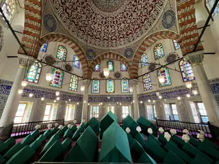 An ornate mosque interior features painted arches, stained-glass windows, hanging lamps, and rows of green-covered tombs.