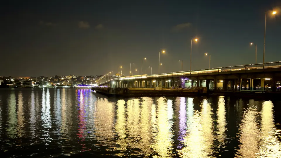 A long illuminated bridge spans dark water, casting bright reflections across the surface at night.