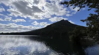 A forested mountain rises above a calm lake reflecting a partly cloudy blue sky.