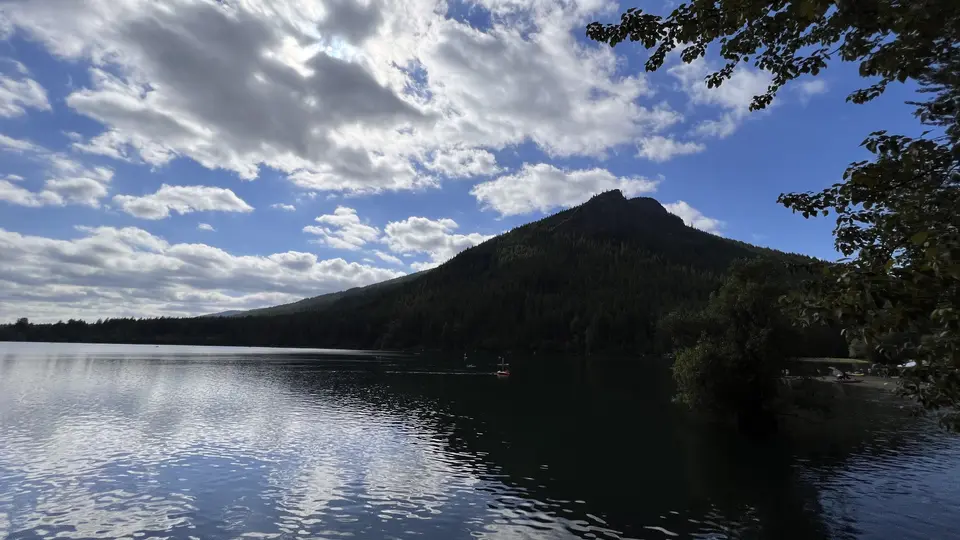 A forested mountain rises above a calm lake reflecting a partly cloudy blue sky.