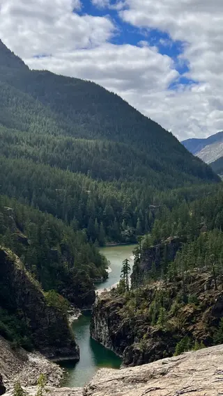 A turquoise river winds through steep rocky cliffs beneath a densely forested mountain and scattered clouds.