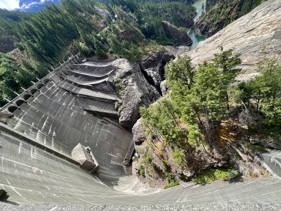 A massive curved concrete dam spans a rocky forested gorge above green water.