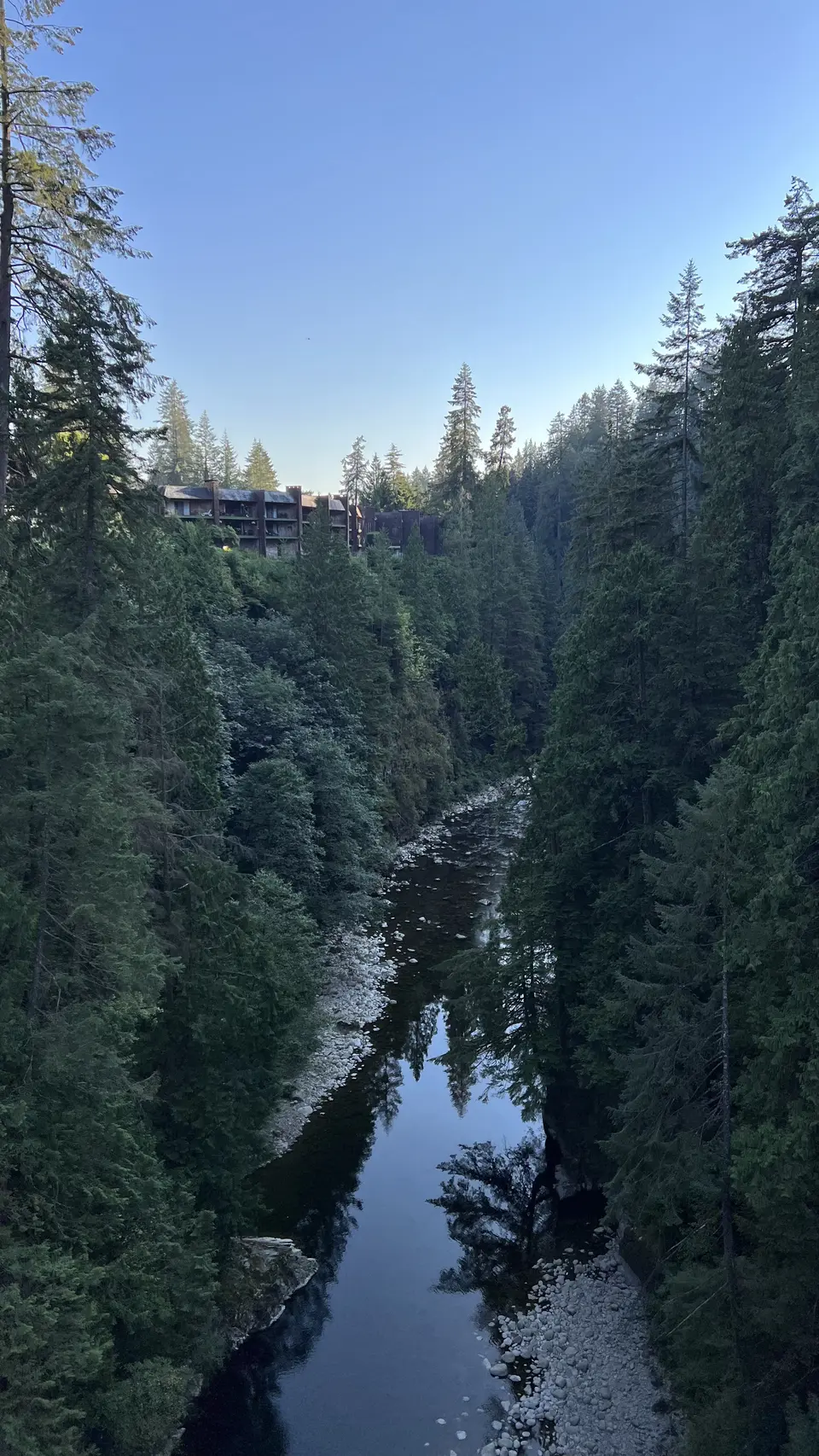 A dark river winds through a steep forested gorge beneath a clear blue sky, with a building partly visible among the trees.