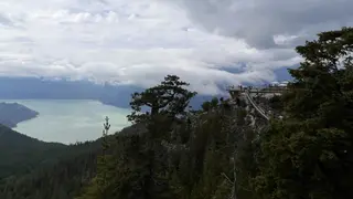 A viewing platform clings to a forested rocky mountainside overlooking a pale lake beneath heavy clouds.