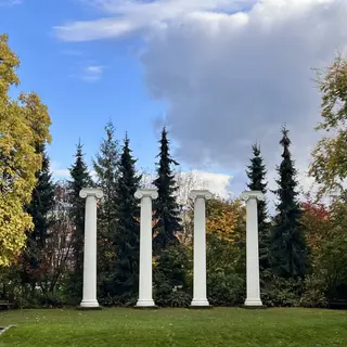 Four white classical columns stand on a lawn before evergreen and autumn-colored trees beneath a cloudy blue sky.