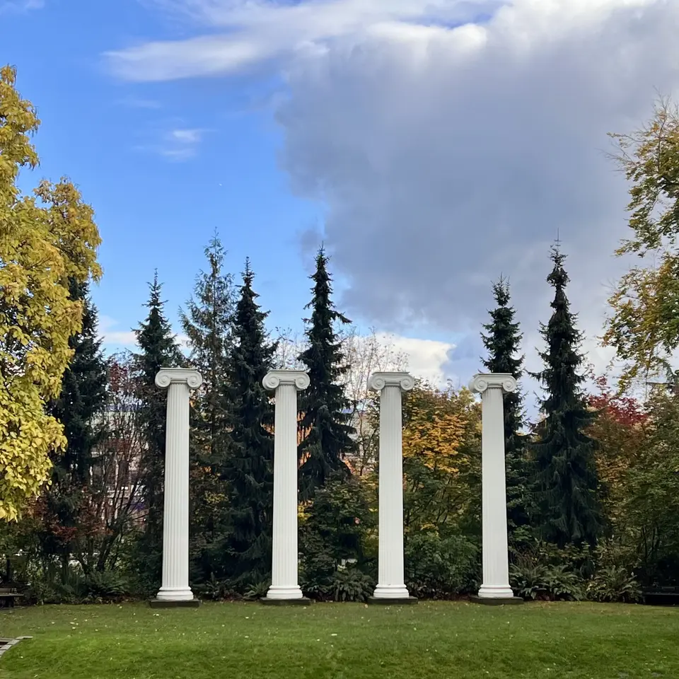Four white classical columns stand on a lawn before evergreen and autumn-colored trees beneath a cloudy blue sky.