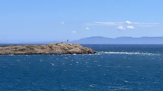 A small rocky island with a beacon rises from choppy blue water beneath a clear sky and distant mountains.