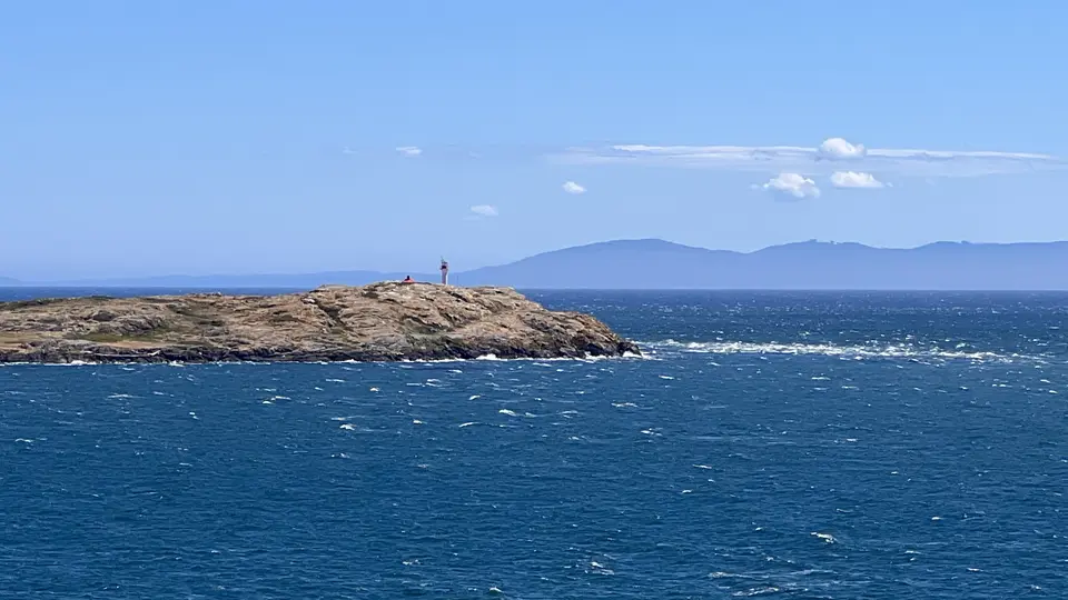 A small rocky island with a beacon rises from choppy blue water beneath a clear sky and distant mountains.