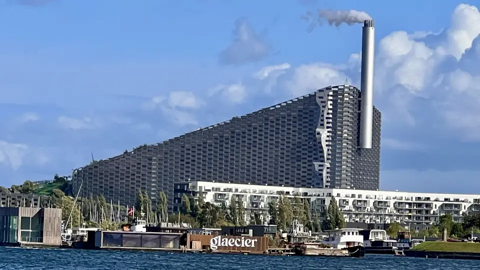 A large modern waterfront building with a tall smokestack rises behind boats and a floating structure marked “glæcier” under a blue sky.