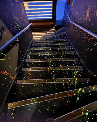A black staircase splattered with bright neon paint descends between dark walls with metal handrails.