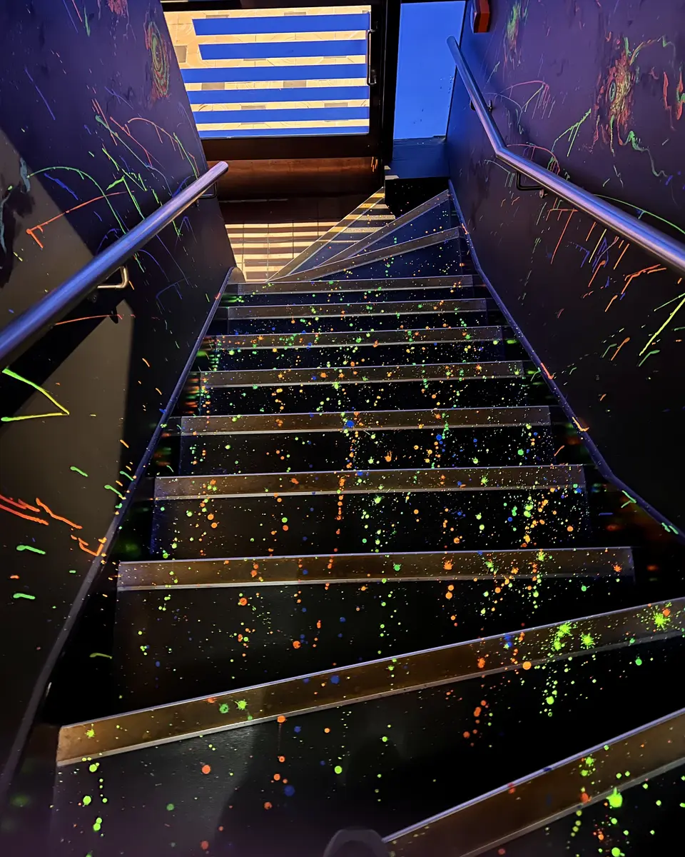A black staircase splattered with bright neon paint descends between dark walls with metal handrails.