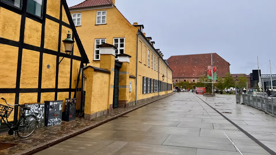 Yellow historic buildings line a wet paved walkway beside a bicycle and a waterfront railing.