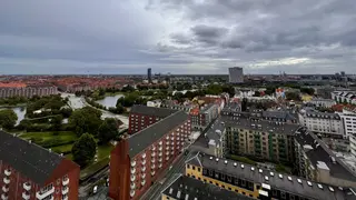 A high-angle view overlooks dense city blocks, a green park, and a small body of water beneath a cloudy sky.