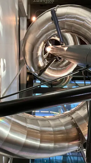 A shiny metal tube slide curves through an indoor structure with black support rails.