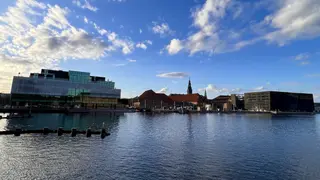 Modern waterfront buildings and a church steeple stand across rippling water beneath a blue sky with scattered clouds.