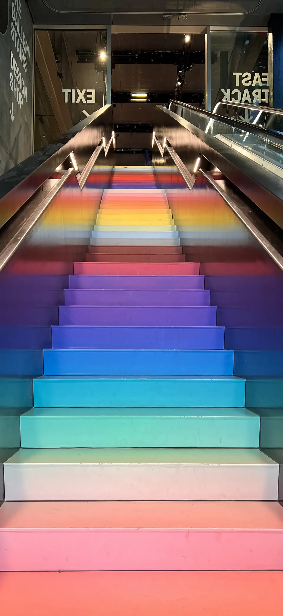 A broad staircase illuminated in rainbow colors rises between metal handrails beside an escalator, with reversed EXIT and EAST TRACK signs visible in glass reflections.