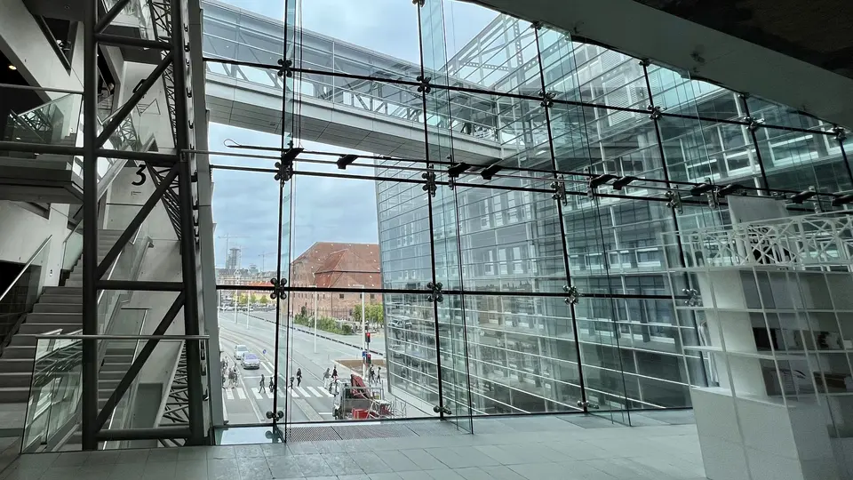 A glass-walled modern interior overlooks a city street and neighboring office buildings through a grid of metal supports.