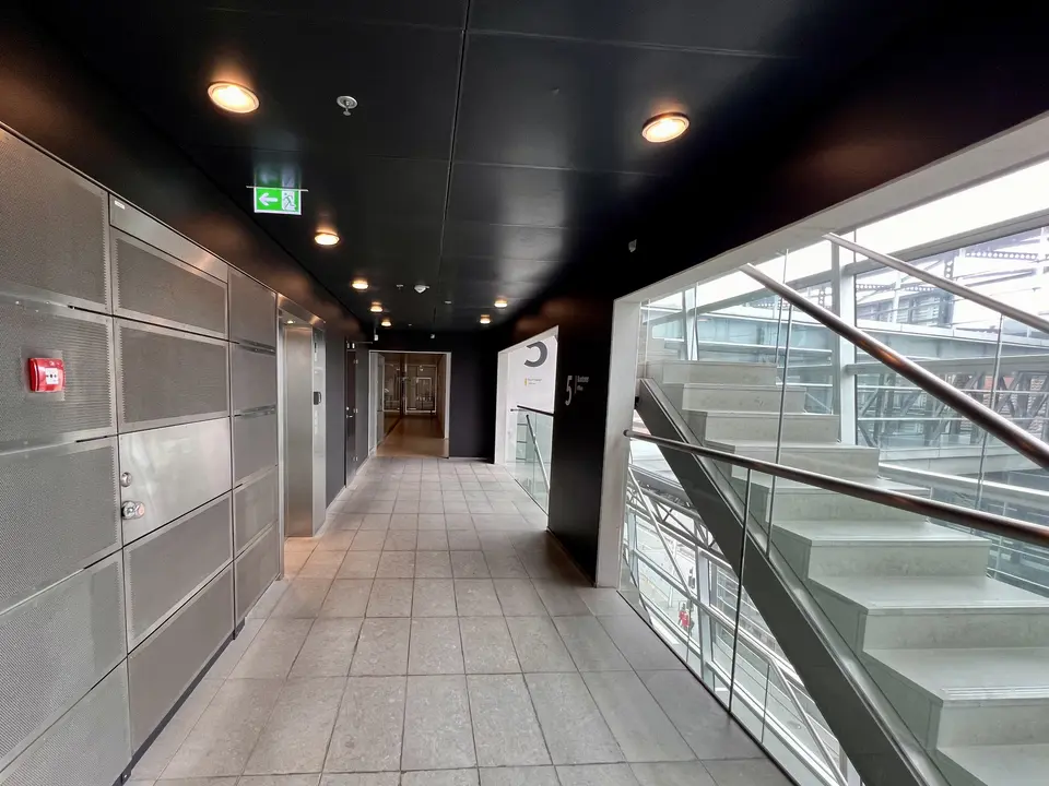 A tiled interior corridor with metal wall panels runs beside a glass-railed staircase under recessed ceiling lights.