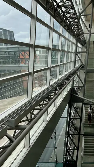 A multi-level interior atrium features large glass windows, steel trusses, and a staircase beside an elevated walkway.