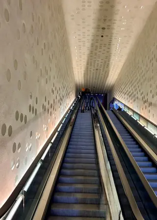 Two parallel escalators descend through a brightly lit corridor with walls and ceiling patterned in circular perforations.