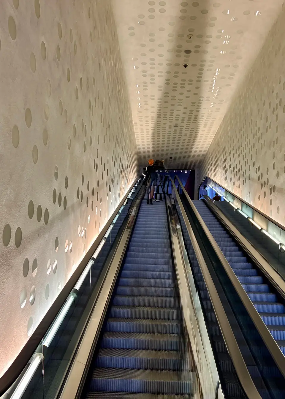 Two parallel escalators descend through a brightly lit corridor with walls and ceiling patterned in circular perforations.