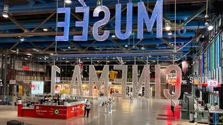 Oversized illuminated letters hang upside down in a spacious transit hall with ticket counters, stairs, and scattered visitors.