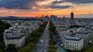 A broad tree-lined avenue leads through a dense cityscape toward a glowing sunset beneath dramatic clouds.