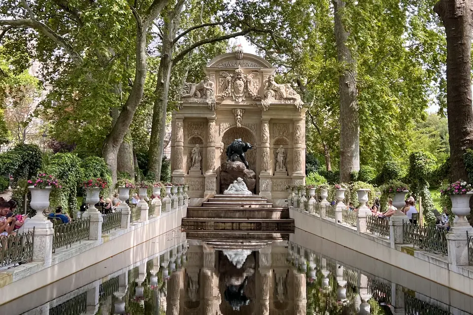 An ornate stone fountain monument with sculpted figures stands at the end of a narrow reflecting pool beneath leafy trees.