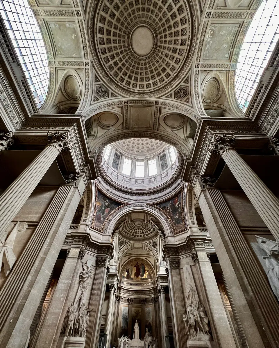 An ornate vaulted interior with coffered ceilings, skylights, and massive fluted columns rises above classical statues.