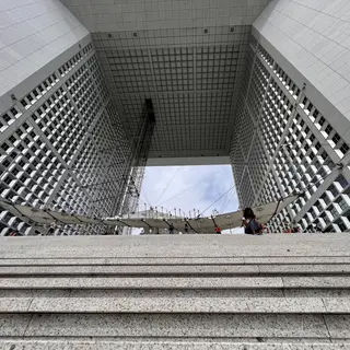 Broad stone steps lead toward the open central arch of a towering grid-windowed building, where people gather beneath a suspended canopy.