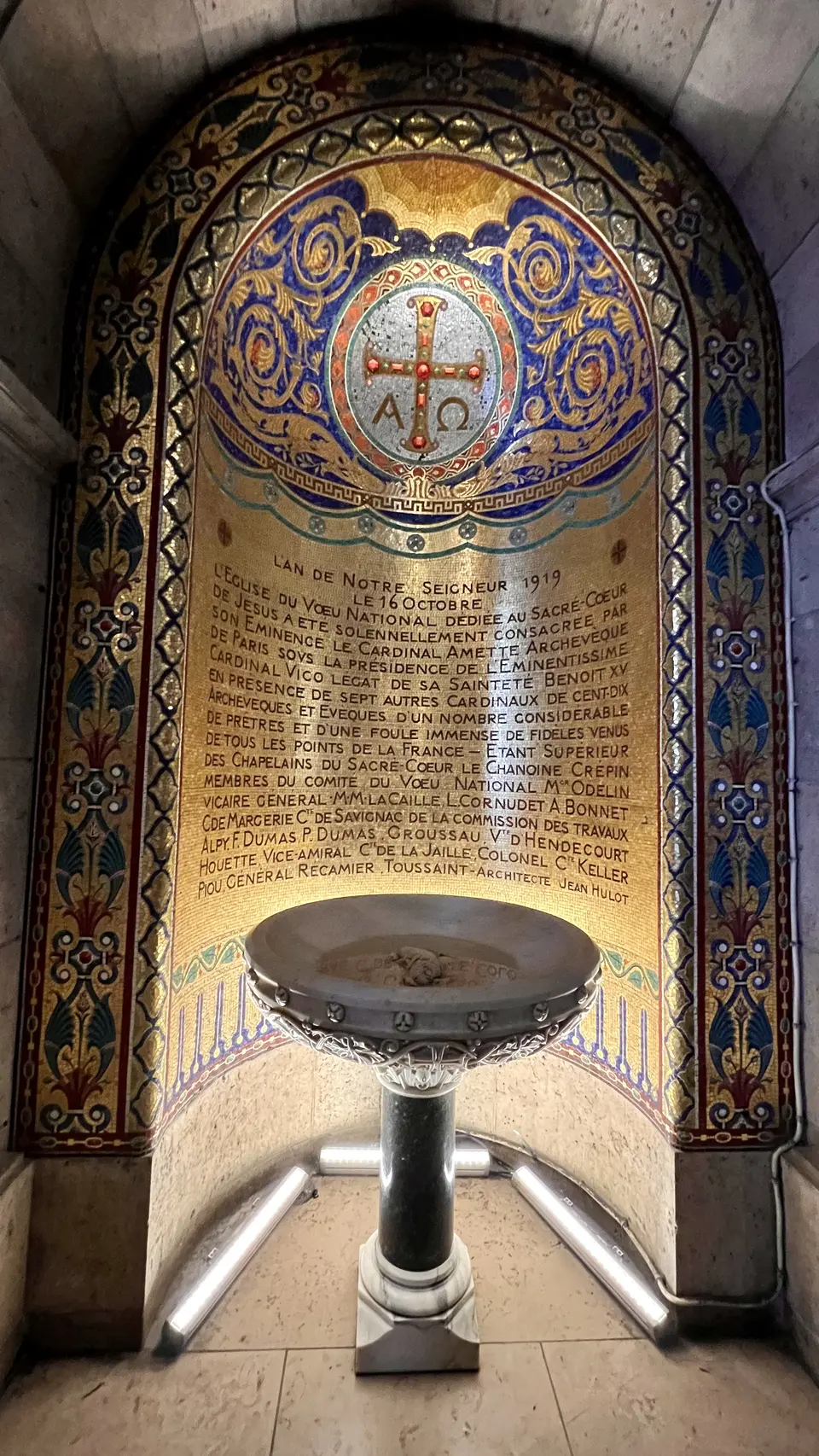 An ornate gold and blue mosaic panel with a cross emblem and French inscription rises behind a decorative stone basin.