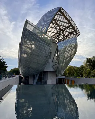 A dramatic curved glass-and-steel building rises above a reflecting pool beneath a lightly clouded sky.
