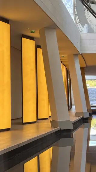 A modern interior corridor features glowing amber wall panels, angled white columns, and reflections in a dark water feature.